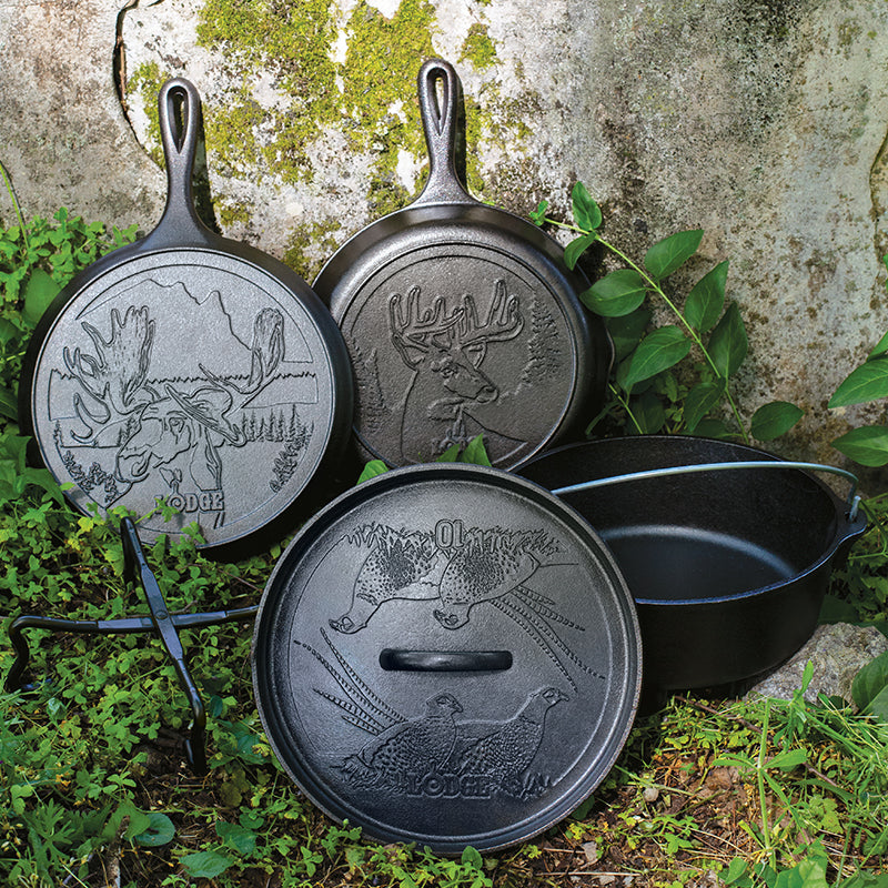 A set of seasoned cast iron cookware including a skillet, griddle, and a camp dutch oven with engraved wildlife designs, displayed on a natural stone background.